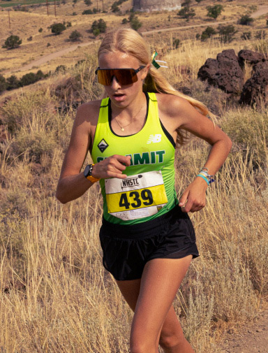 A young female athlete is running in an outdoor cross-country race. She is wearing a lime green and black "SUMMIT" athletic top with the number "439" pinned to it, black shorts, and large, wrap-around sunglasses. Her blonde hair is tied back with a green ribbon, and she appears focused and determined as she runs through a dry, grassy terrain with scattered bushes and rocks. The landscape in the background is arid and hilly, indicative of a challenging race environment.