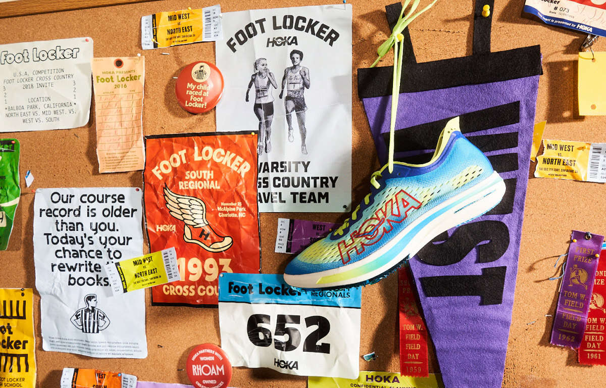 A bulletin board filled with various memorabilia related to Foot Locker cross-country events. The items include several posters, race bibs, ticket stubs, and ribbons. Prominent among them is a blue and yellow HOKA running shoe hanging by its laces. There is a vintage Foot Locker South Regional poster from 1993, a flyer with the text "Our course record is older than you. Today’s your chance to rewrite the books," and a "Foot Locker Varsity Cross Country Travel Team" poster with images of runners. The board is a vibrant collage celebrating past races and achievements.