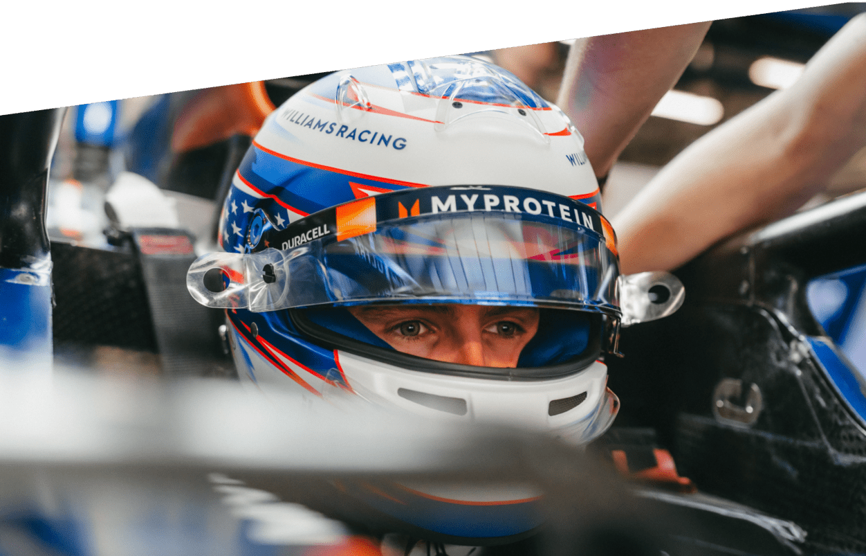 Close-up of a Formula 1 driver wearing a helmet and seated in the Williams Racing car. The helmet features branding from Duracell. The driver has a focused expression, with eyes visible through the visor, while team members are working around the car in the background.