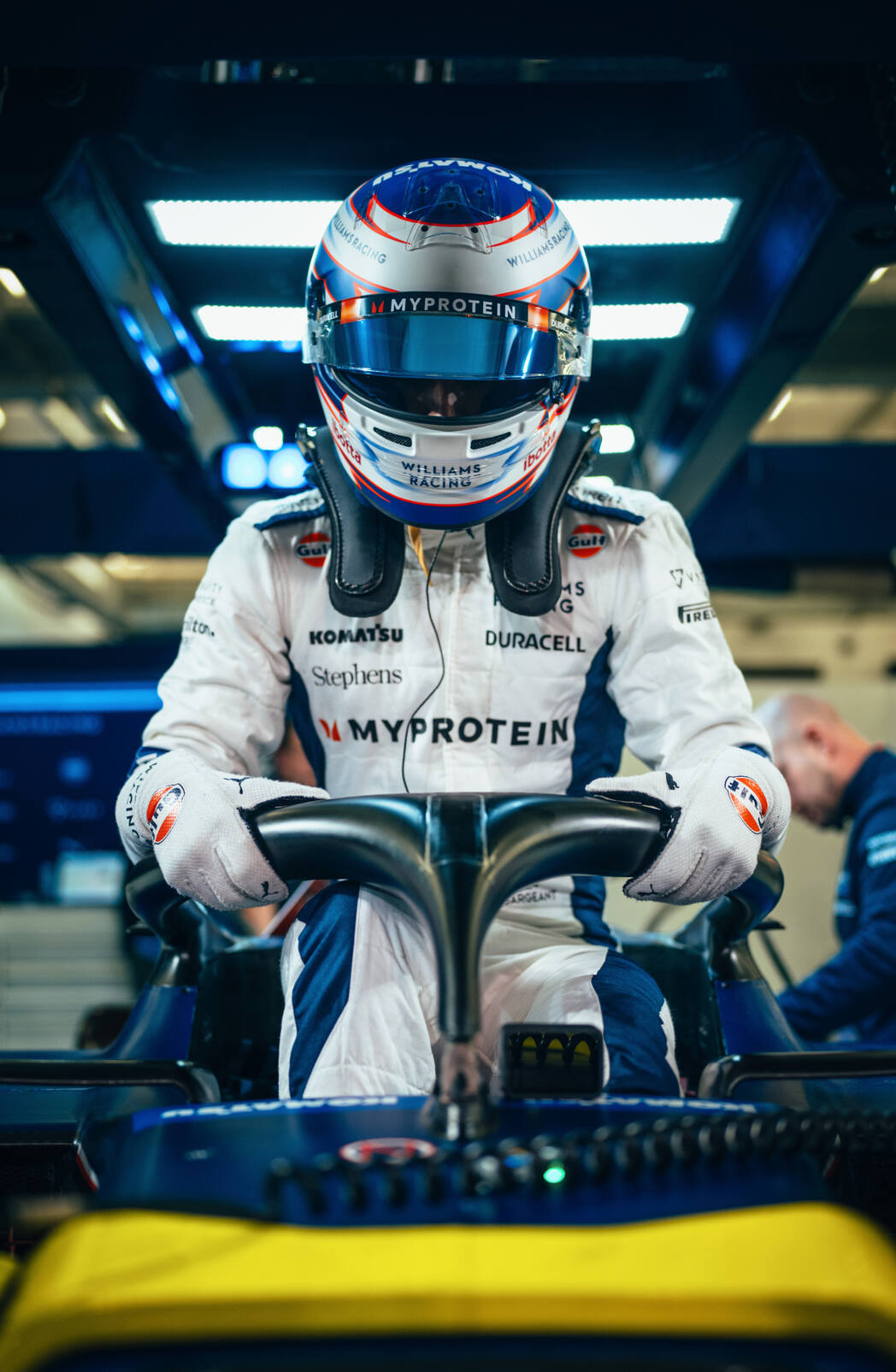 Front view of a Formula 1 driver in a Williams Racing suit and helmet, standing behind the steering wheel of a racing car. The driver's helmet and suit features branding from Duracell. The driver appears to be focused, with a technician visible in the background working in the garage.