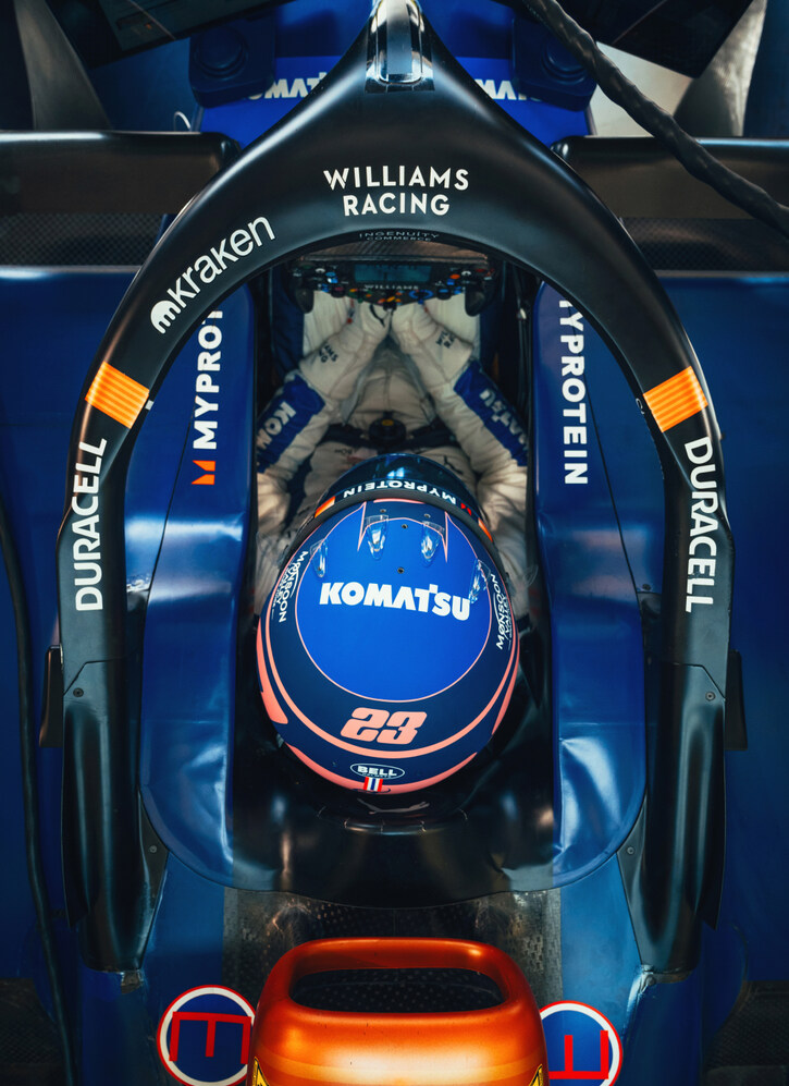 Overhead view of a Formula 1 driver in a Williams Racing car cockpit. The driver is wearing a blue helmet with the number 23 and the logo of Komatsu. The car features prominent branding from Duracell on the halo and bodywork. The scene captures the driver in a focused moment, with their hands on the steering wheel and the detailed design of the car's interior visible.