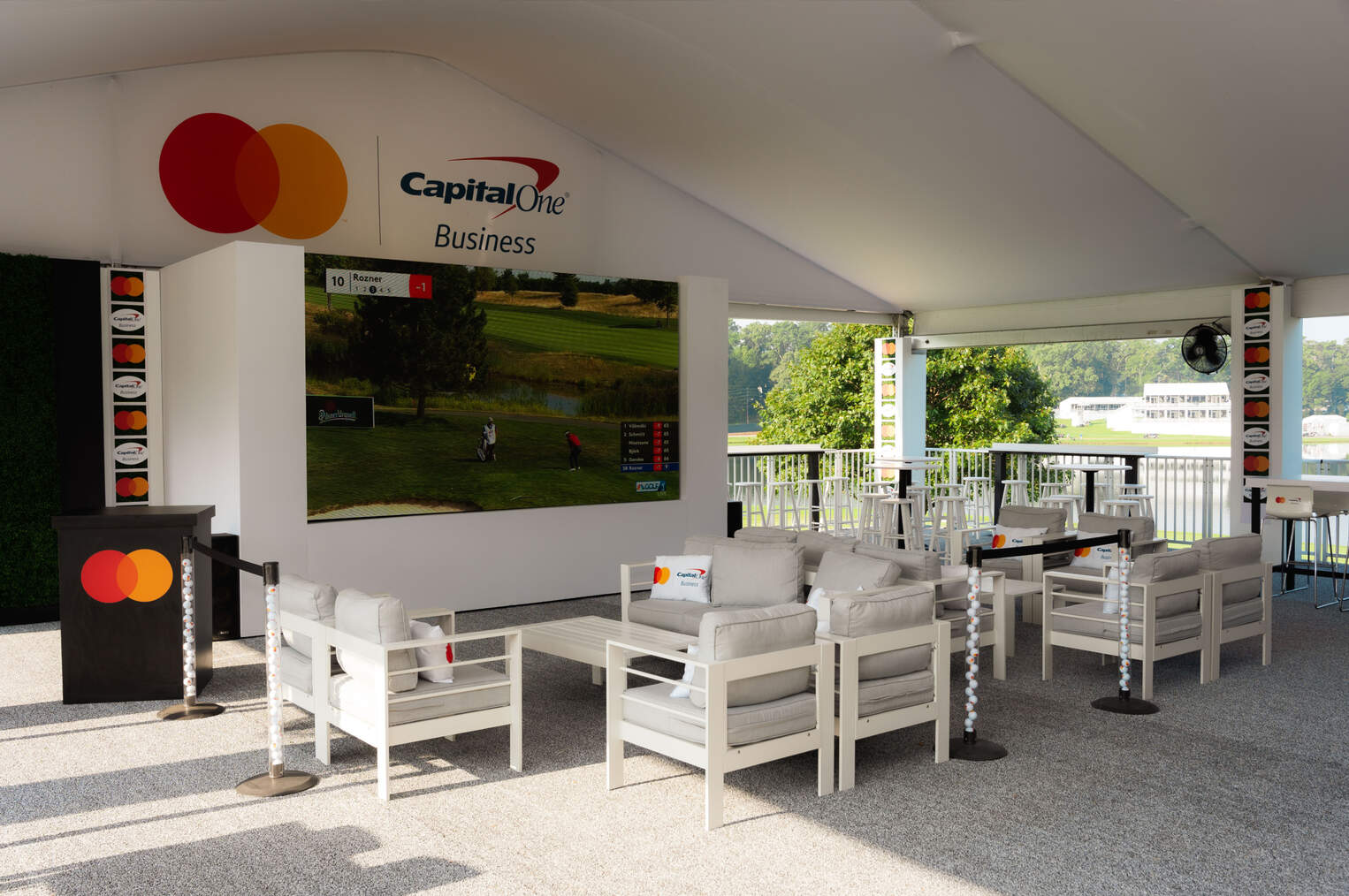 Capital One Business seating area with white furniture, a large screen showing a golf course, and branded decorations under a canopy.