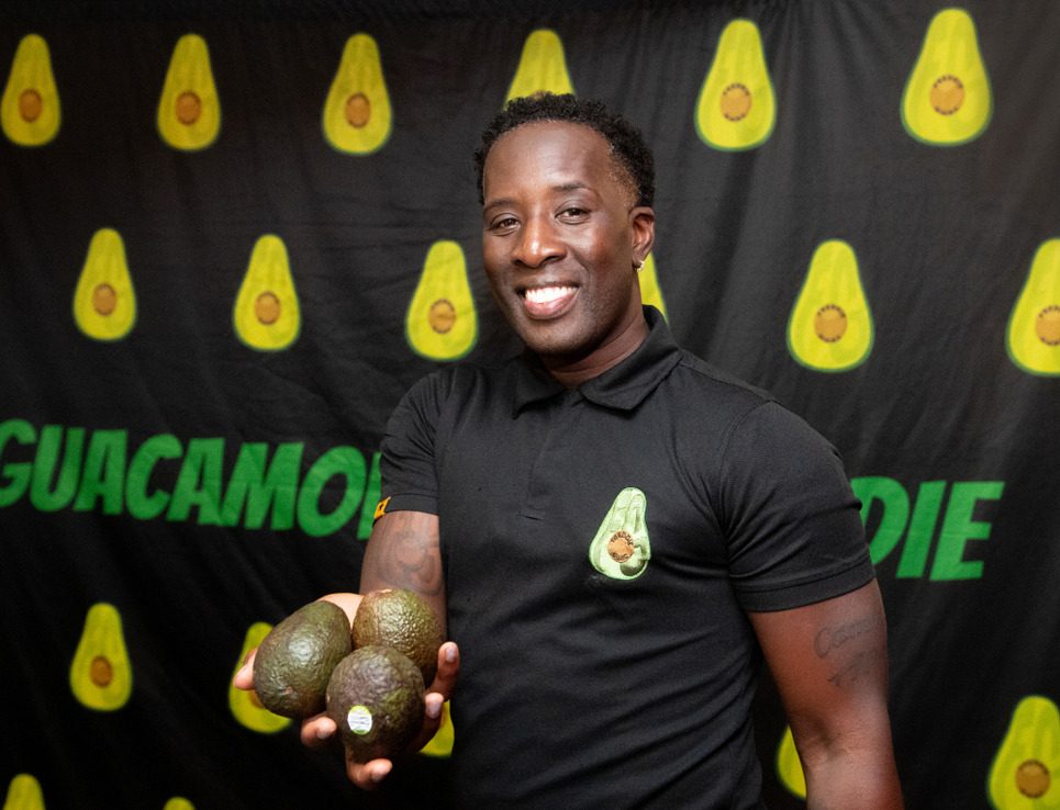 Man holding avocados in front of a backdrop with avocado graphics and the word 'Guacamole' wearing a black shirt with an avocado logo.