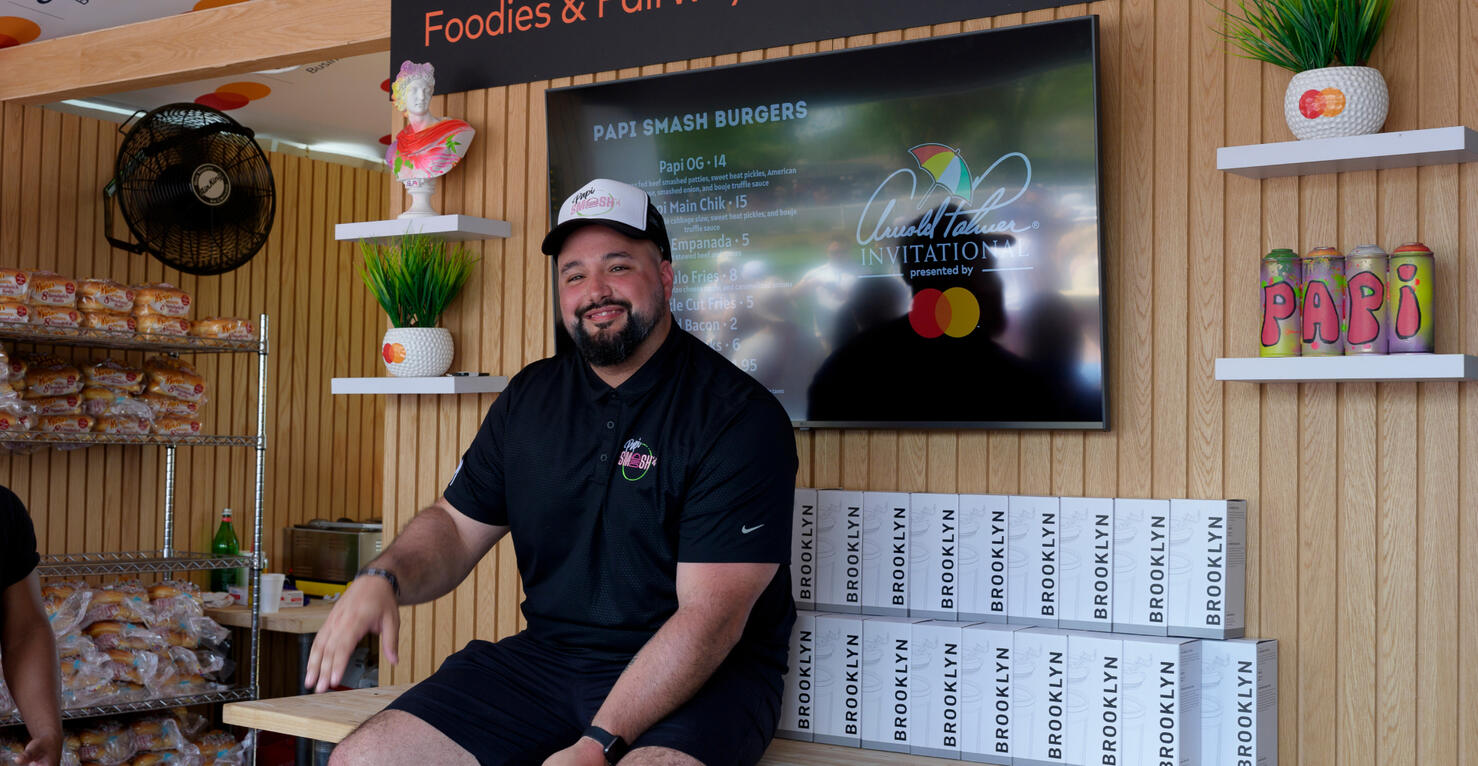 Man seated in a food stall wearing a 'Papi Smash' polo, with a screen displaying the menu and the 'Arnold Palmer Invitational' logo behind him.