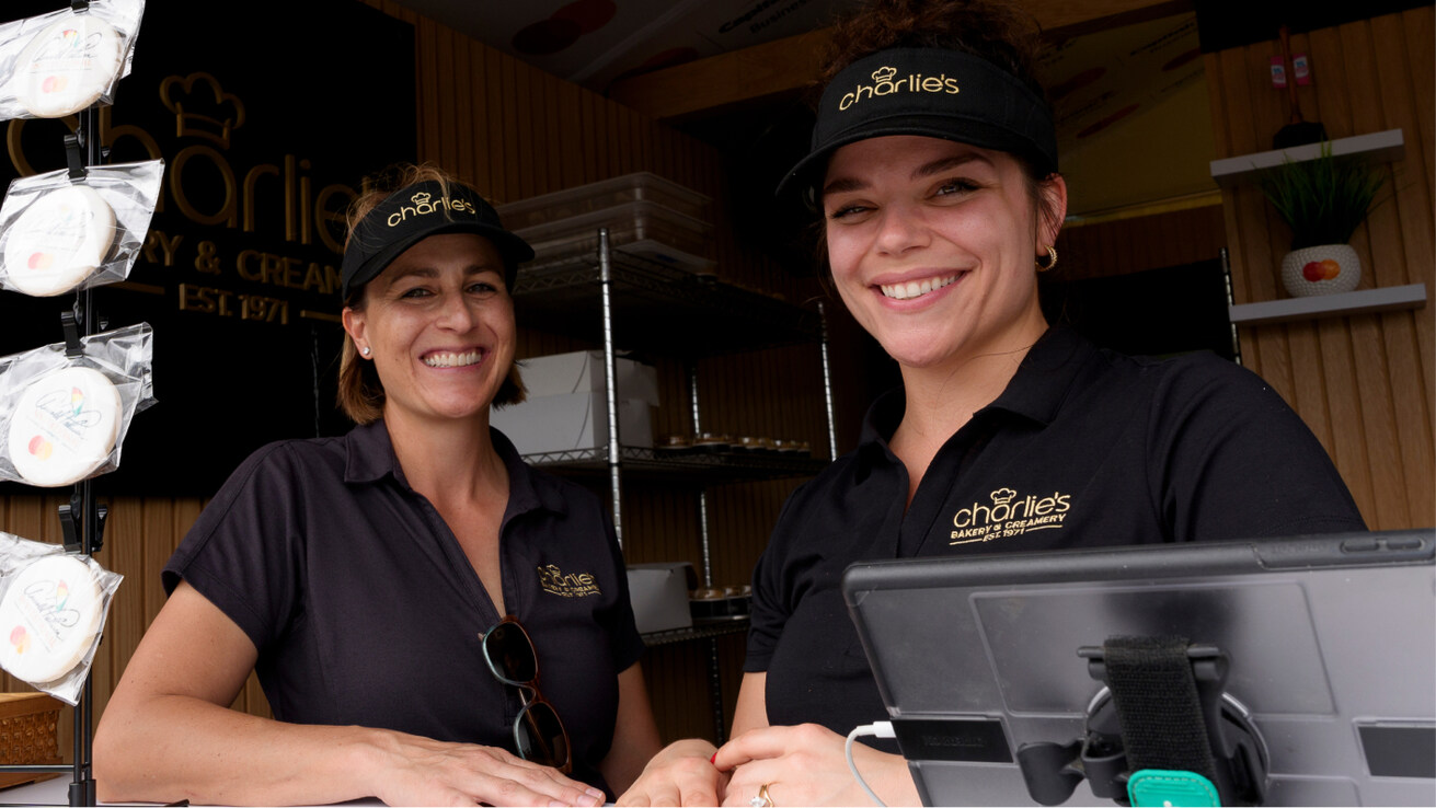 Two smiling women in black visors and shirts at Charlie's Bakery & Creamery stand, with cookies and a logo sign in the background.