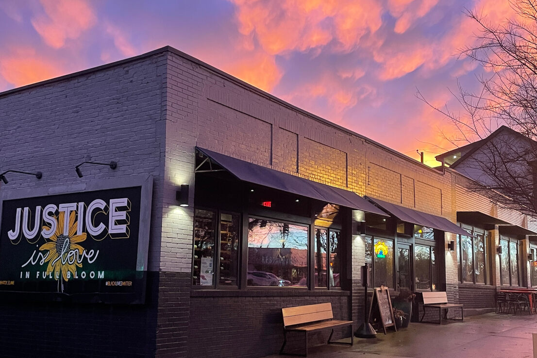 Brick building at sunset with a sign reading 'Justice is Love in Full Bloom' and a flower graphic, with a colorful sky in the background.