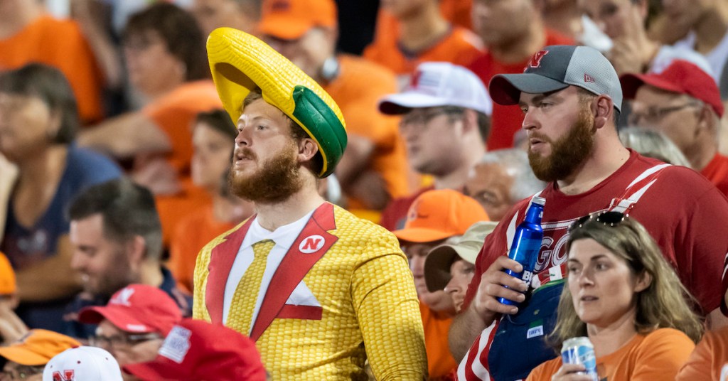 A Cornhuskers Fan watches a Nebraska football game while wearing a hat shaped like corn on the cob.