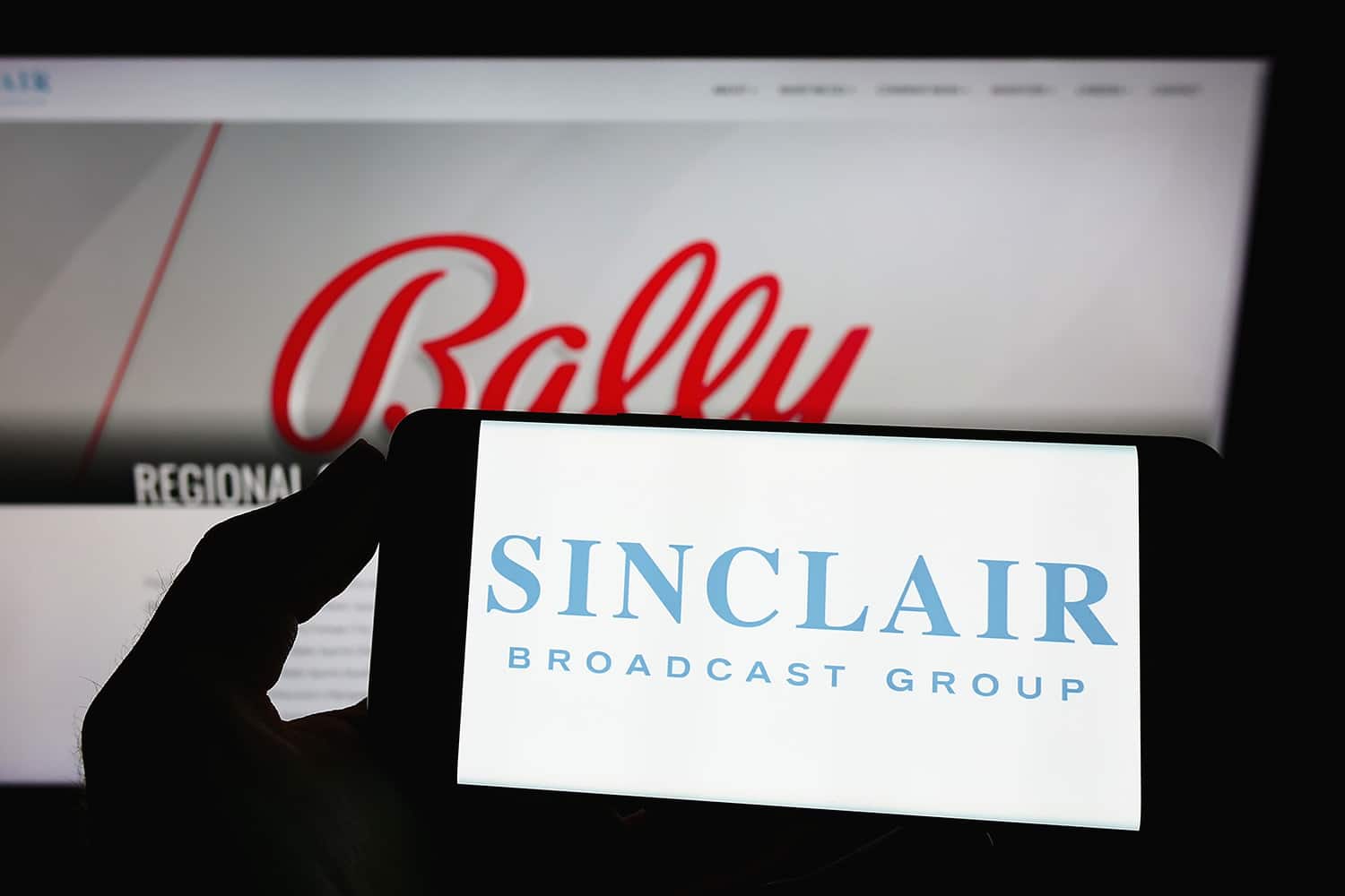 A phone displaying the Sinclair Broadcast Group logo is shown in front of the logo of their RSN brand, Bally Sports.