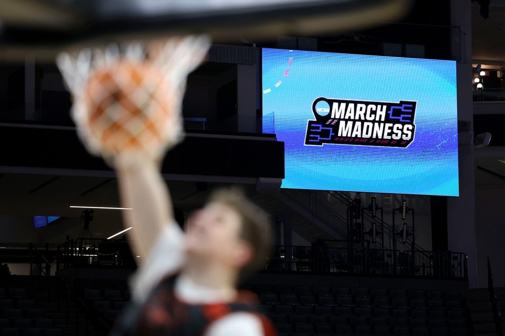 A basketball player dunks a basketball with the March Madness logo on a big screen prominently in the background.