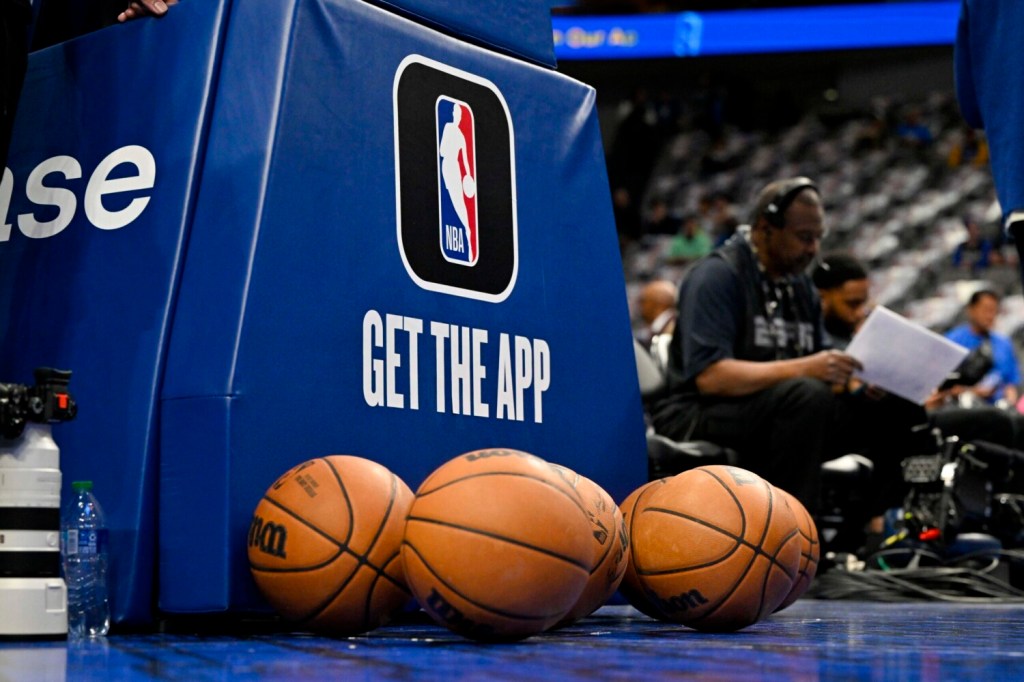 Basketballs gathered at the stanchion of an NBA basket, which is advertising the NBA app.