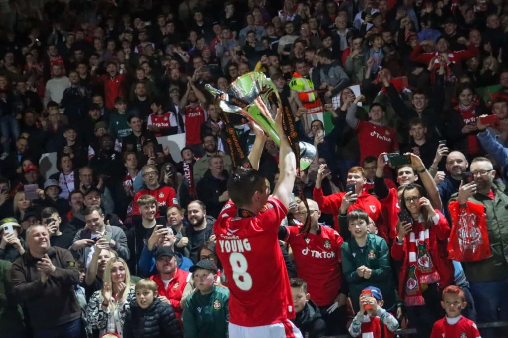 Wrexham AFC player Luke Young lifts the National League trophy in front of fans.