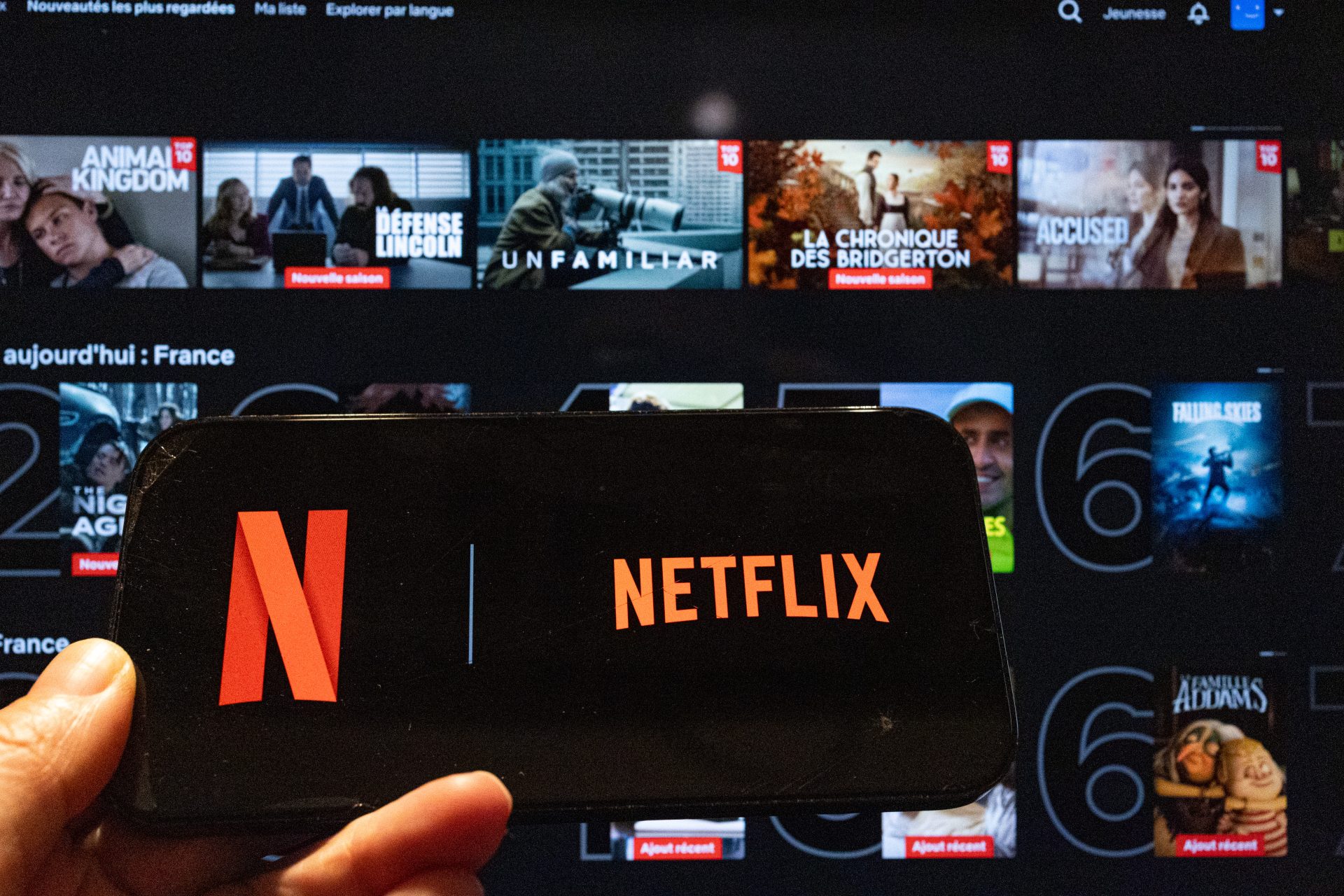 A smartphone showing the Netflix logo is held in front of a television displaying the Netflix home screen with Top 10 content rows in Paris, Ile de France, France, February 28, 2026. The scene illustrates video on demand streaming and second screen viewing on connected devices.
